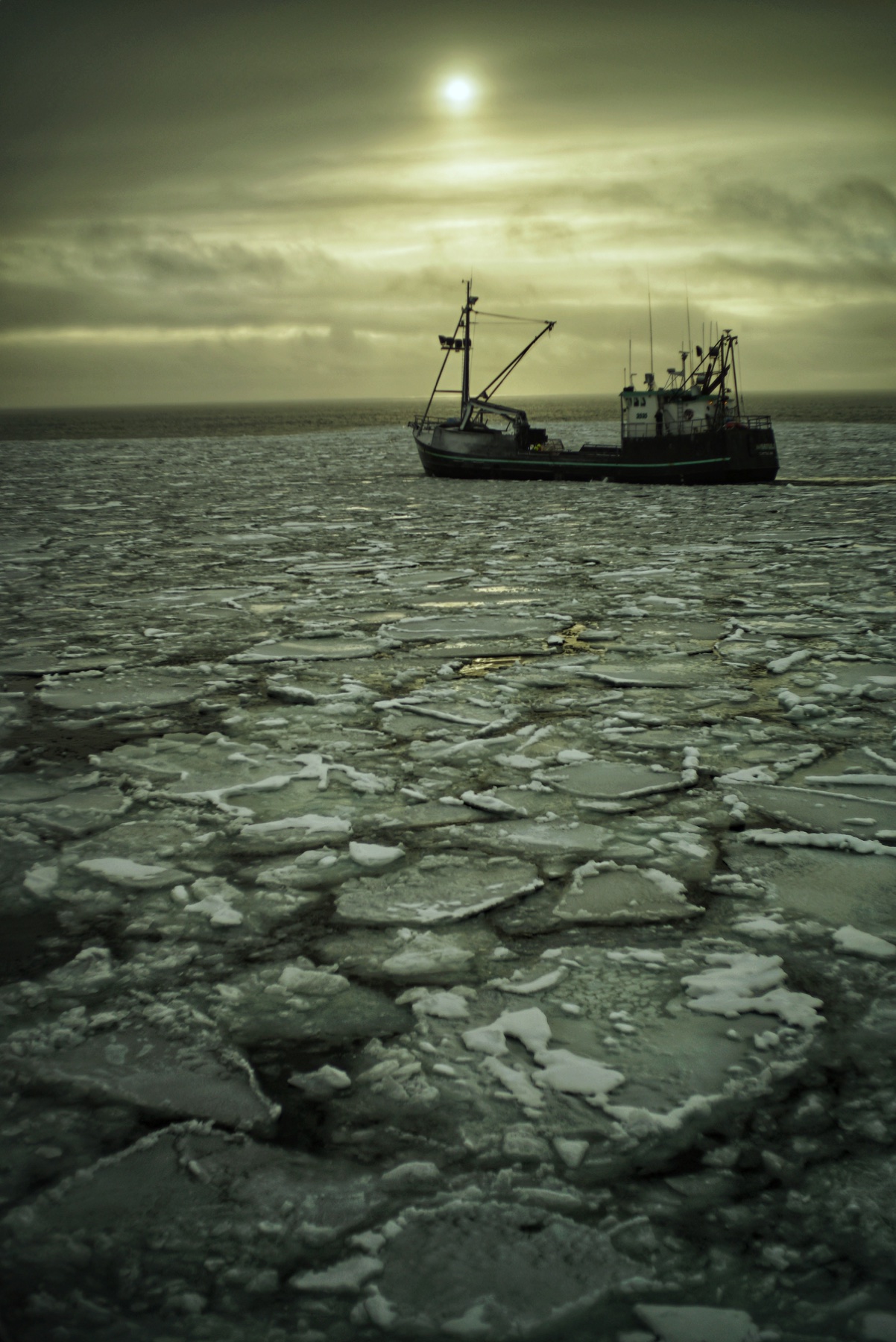 A fishing vessel moving through broken ice beneath a low winter sun