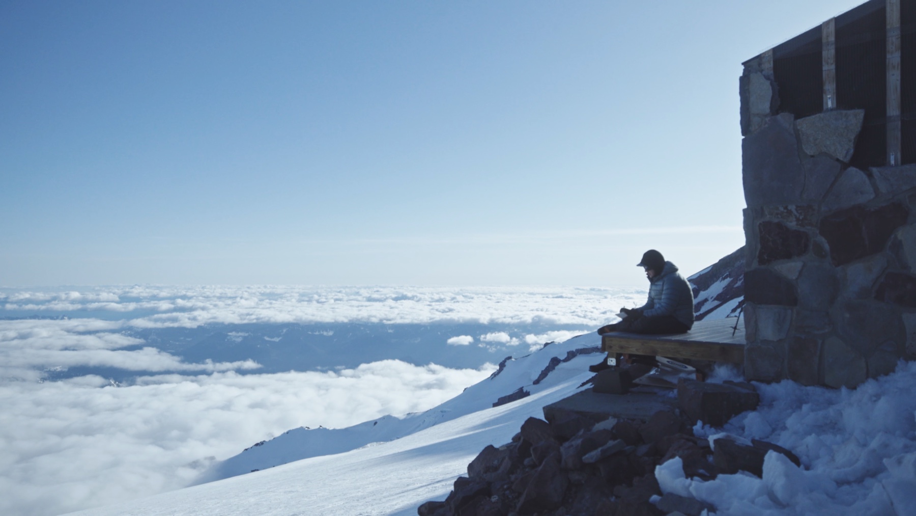 A crew member seated beside a mountain hut above the cloud line